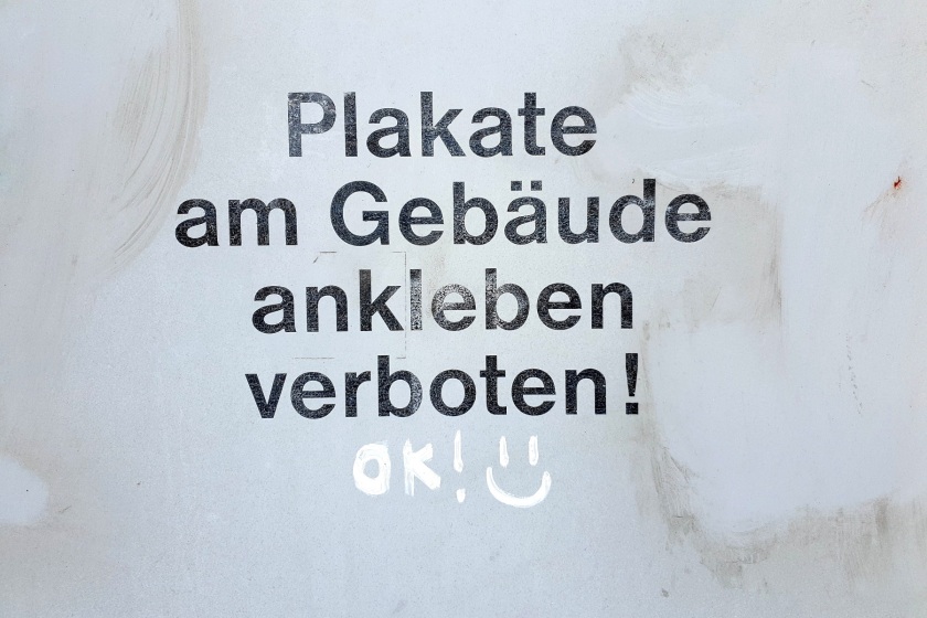 Graue Fläche mit der gedruckten Aufschrift "Plakate am Gebäude ankleben verboten!", darunter handschriftlich "OK! :)"